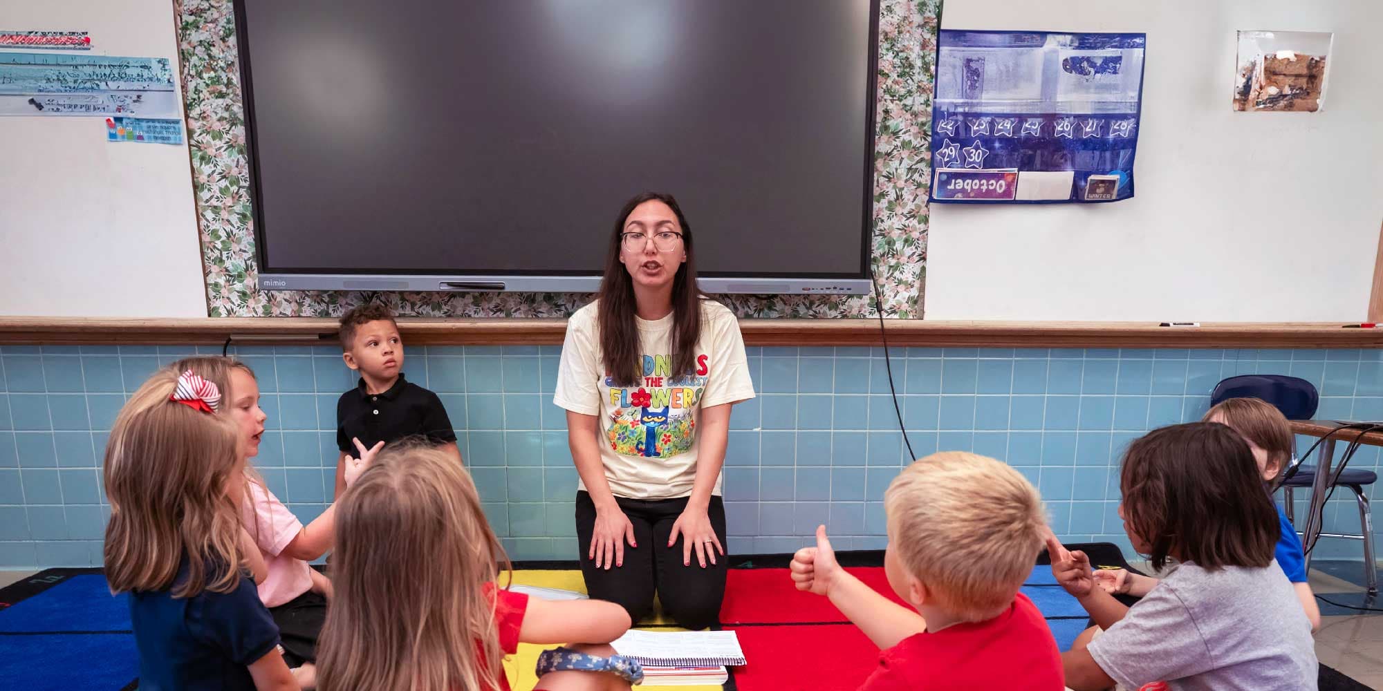 Teacher kneeling in front of a classroom screen, speaking with students seated on the floor.